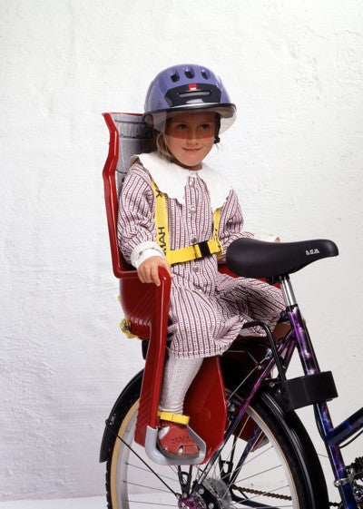 A child wearing a purple bicycle helmet sits in a red and yellow bicycle child seat mounted on a purple bicycle against a white textured wall.